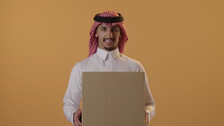عملية توصيل الطلبات للزبائن، التسوق وطلب المنتجات عبر الإنترنت، خدمات الشحن و التوصيل ذات الجودة العالية، بورتريه لشاب عربي خليجي سعودي يرتدي الثوب و الشماغ يحمل بيده صندوق كرتوني، خلفية برتقالية