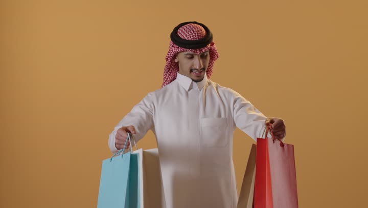 المشتريات التجارية والهدايا، التبضع وأخذ الحاجات اللازمة, مواسم التخفيضات في السعودية, الاستفادة من الحسومات والتصفيات, بورتريه لشاب عربي خليجي سعودي يرتدي الثوب التقليدي والشماغ يمسك بيده أكياس ورقية ملونة, متعة التسوق في المملكة, خلفية برتقالية