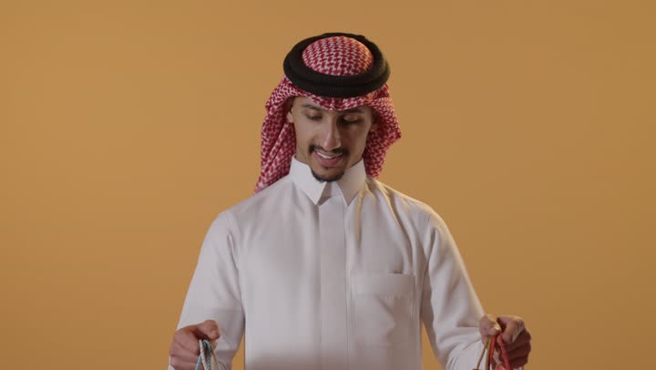 التبضع وأخذ الحاجات اللازمة, المشتريات التجارية والهدايا, مواسم التخفيضات في السعودية, الاستفادة من الحسومات والتصفيات, بورتريه لشاب عربي خليجي سعودي يرتدي الثوب التقليدي والشماغ يمسك بيده أكياس ورقية ملونة, متعة التسوق في المملكة, خلفية برتقالية