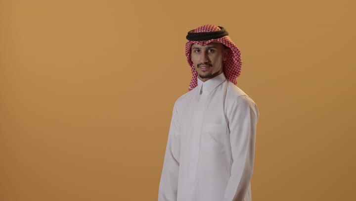 الوقوف بايدي متقاطعة ، مفهوم الرجولة والقوة والثقة بالنفس، بورتريه لرجل عربي خليجي سعودي يرتدي الشماغ والزي التقليدي ينظر الى الكاميرا بإيماءات السرور والسعادة ، الاهتمام بالأناقة والمظهر الخارجي ، خلفية برتقالية