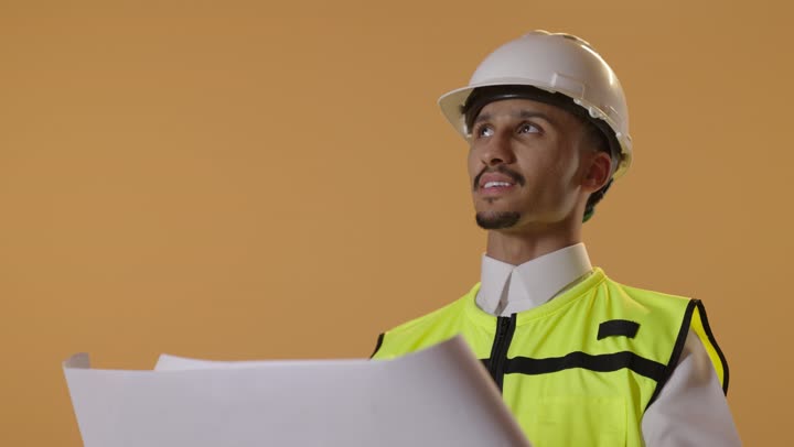 مفهوم ادارة المشاريع وتدقيقها، العمل في القطاع الهندسي، بورتريه مقرب  لمهندس عربي خليجي سعودي يرتدي الزي التقليدي مع سترة الحمايه والخوذة يقوم بدراسة المخططات الهندسية، خلفية برتقالية