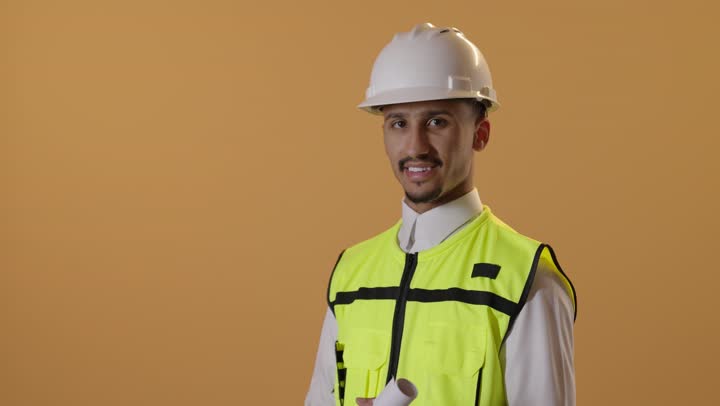 متابعة سير المشاريع الإعمارية، مفهوم الإنشاءات المعمارية والهندسة، بورتريه لمهندس عربي خليجي سعودي يرتدي الثوب الابيض والخوذة وسترة الحماية يحمل بيده مخطط ينظر إلى الكاميرا بإيماءات الثقة، المهن والوظائف الشبابية، خلفية برتقالية