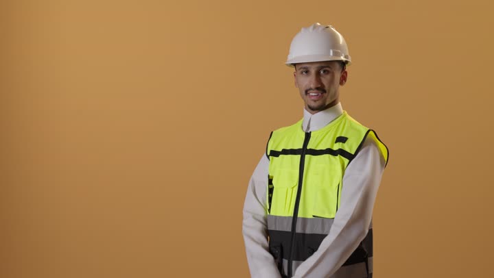 مهن ووظائف سعودية, بورتريه لرجل عربي سعودي خليجي يرتدي الثوب والسترة ويضع خوذه الحماية يقف بايدي متقاطعة بتعابير السعادة والسرور, خلفية برتقالية