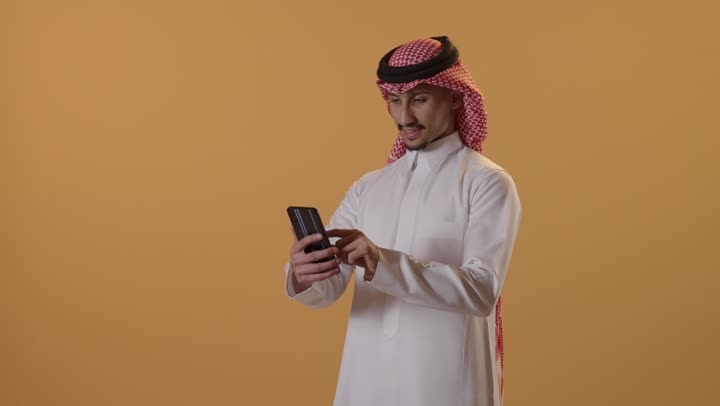 Browsing the internet and online shopping, using modern technologies and devices, communicating with individuals via mobile phones, chatting and conversations and meeting new people, a portrait of a smiling Arab Gulf Saudi man wearing a white thobe and a ghutrah using a mobile phone, with an orange background.