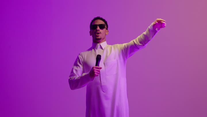 تقديم العروض الصوتية،  تعابير الاندماج والتفاعل مع الموسيقا، تسجيل مقطع صوتي غنائي، بورتريه لشاب عربي خليجي سعودي مبتسم يرتدي الثوب الابيض و النظارة الشمسية يمسك بيده المايكروفون يقوم بالغناء بإيماءات الفرح والسرور، خلفية ملونه