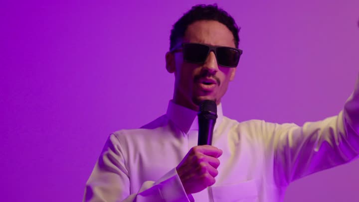 تسجيل مقطع صوتي غنائي، تقديم العروض الصوتية،  تعابير الاندماج والتفاعل مع الموسيقا، بورتريه مقرب لشاب عربي خليجي سعودي مبتسم يرتدي الثوب الابيض و النظارة الشمسية يمسك بيده المايكروفون يقوم بالغناء بإيماءات الفرح والسرور، خلفية ملونه