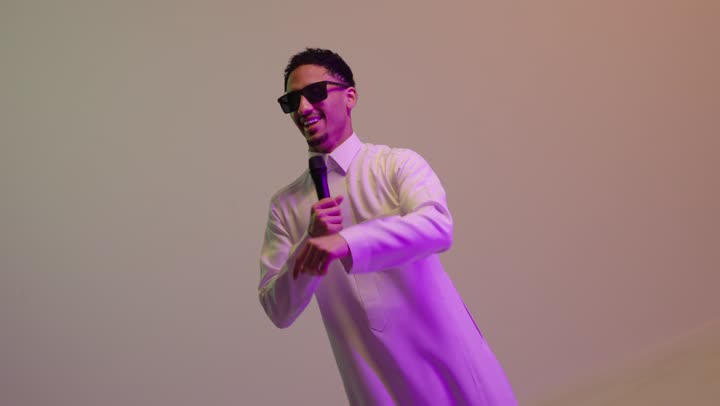 إيماءات الفرح والسرور، تسجيل مقطع صوتي غنائي، تقديم العروض الصوتية،  تعابير الاندماج والتفاعل مع الموسيقا، بورتريه لشاب عربي خليجي سعودي مبتسم يرتدي الثوب الابيض و النظارة الشمسية يمسك بيده المايكروفون يقوم بالغناء، خلفية ملونه