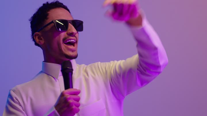 تعابير الاندماج والتفاعل مع الموسيقا، تقديم العروض الصوتية، تسجيل مقطع صوتي غنائي، بورتريه مقرب لشاب عربي خليجي سعودي مبتسم يرتدي الثوب الابيض و النظارة الشمسية يمسك بيده المايكروفون يقوم بالغناء بإيماءات الفرح والسرور، خلفية ملونه