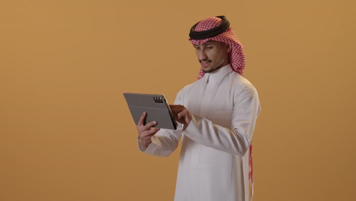 استخدام تطبيقات وبرامج الأجهزة اللوحية، تصفح مواقع التواصل الاجتماعي، الدردشة مع الأهل والأصدقاء، استعمال التكنولوجيا والتقنيات الحديثة، بورتريه مقرب لرجل عربي خليجي عماني يرتدي الدشداشة و الكمة يحمل بيده جهاز التابلت، خلفية برتقالية