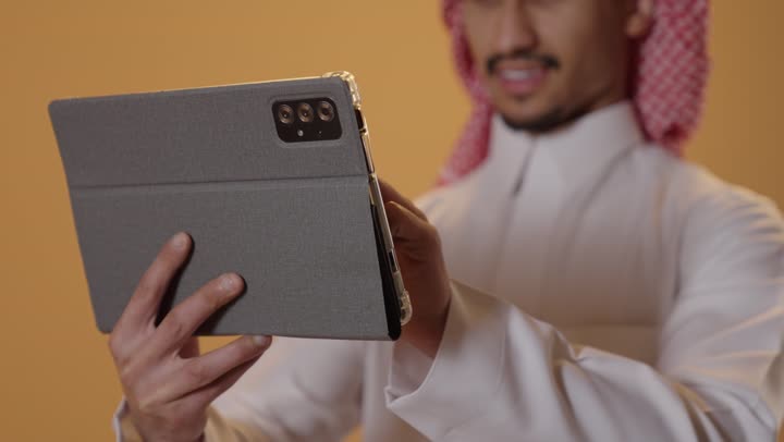 تصفح مواقع التواصل الاجتماعي، استخدام تطبيقات وبرامج الأجهزة اللوحية، الدردشة مع الأهل والأصدقاء، استعمال التكنولوجيا والتقنيات الحديثة، بورتريه مقرب لرجل عربي خليجي عماني يرتدي الدشداشة و الكمة يحمل بيده جهاز التابلت، خلفية برتقالية
