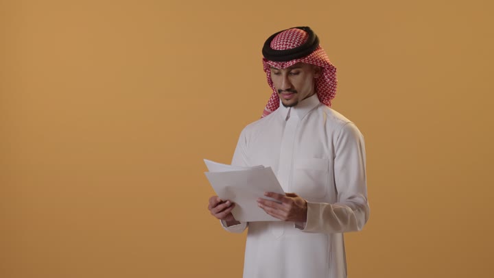 التدقيق في قراءة الملاحظات، اعداد التقارير الخاصة بالعمل، بورتريه لرجل عربي خليجي سعودي الثوب الابيض و الشماغ يحمل بيده مجموعة من الاوراق البيضاء، مراجعة ومتابعة الخطط والمشاريع المكتبية بإيماءات التركيز، خلفية برتقالية