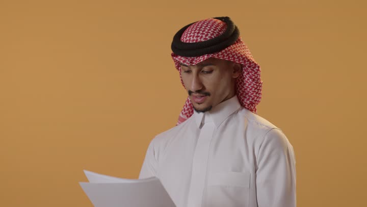 اعداد التقارير الخاصة بالعمل، التدقيق في قراءة الملاحظات، بورتريه مقرب لرجل عربي خليجي سعودي الثوب الابيض و الشماغ يحمل بيده مجموعة من الاوراق البيضاء، مراجعة ومتابعة الخطط والمشاريع المكتبية بإيماءات التركيز، خلفية برتقالية