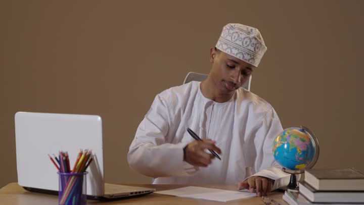 Omani Student Writing at Desk with Laptop and Globe
