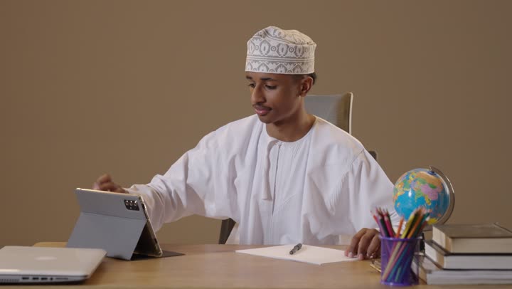 Omani Man Writing and Studying at Desk with Tablet