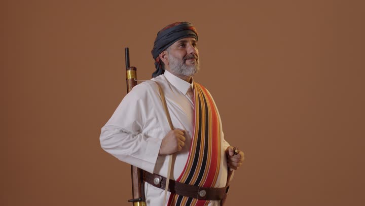 ذكرى تأسيس الدولة السعودية الأولى 22 فبراير، ارتداء أزياء المنطقة الجنوبية للاحتفال في المناسبات الوطنية،  يوم بدينا 1727م، بورتريه  لرجل عربي خليجي سعودي مبتسم يرتدي الثوب والغترة مع الشال الجنوبي يحمل بيده البندقية، خلفية بيج