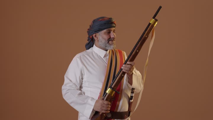 Wearing the traditional attire of the southern region to celebrate national occasions, on the day we began in 1727, a portrait of a smiling Saudi Arabian Gulf man dressed in a thobe and ghutrah with a southern shawl holding a rifle in his hand, commemorating the founding of the first Saudi state on February 22, beige background.