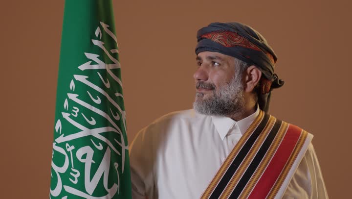 يوم بدينا 1727م، ذكرى تأسيس الدولة السعودية الأولى 22 فبراير، الفخر والاعتزاز بإنجازات الوطن، ارتداء أزياء المنطقة الجنوبية للاحتفال في المناسبات الوطنية، بورتريه  لرجل كبير بالسن عربي خليجي سعودي  يرتدي الثوب والغترة مع الشال الجنوبي يحمل بيده علم التأسيس السعودي ،  خلفية بيج