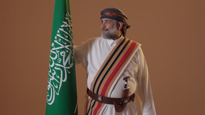 The anniversary of the establishment of the first Saudi state is on February 22, a source of pride and honor in the nation's achievements. Wearing traditional attire from the southern region to celebrate national occasions, the day we began in 1727 AD. A portrait of an elderly Arab Gulf Saudi man wearing a thobe and ghutrah with a southern shawl, holding the Saudi founding flag in his hand, with a beige background.