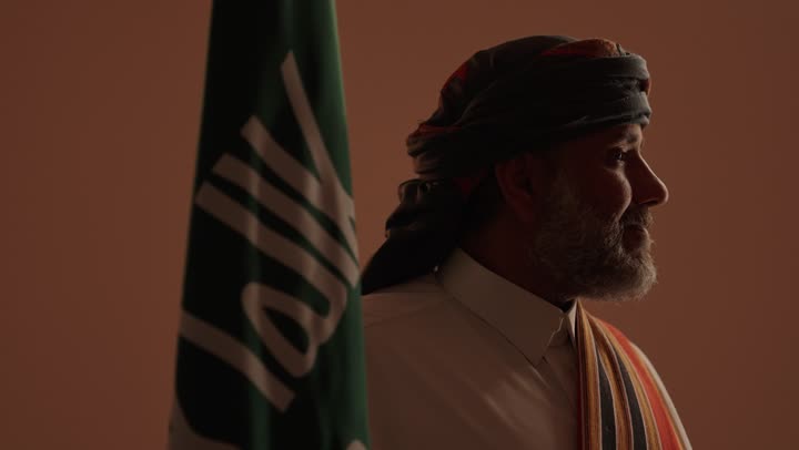 The anniversary of the establishment of the first Saudi state is on February 22, a source of pride and honor in the nation's achievements. Wearing traditional attire from the southern region to celebrate national occasions, the day we began in 1727 AD. A portrait of an elderly Arab Gulf Saudi man wearing a thobe and ghutrah with a southern shawl, holding the Saudi founding flag in his hand, with a beige background.