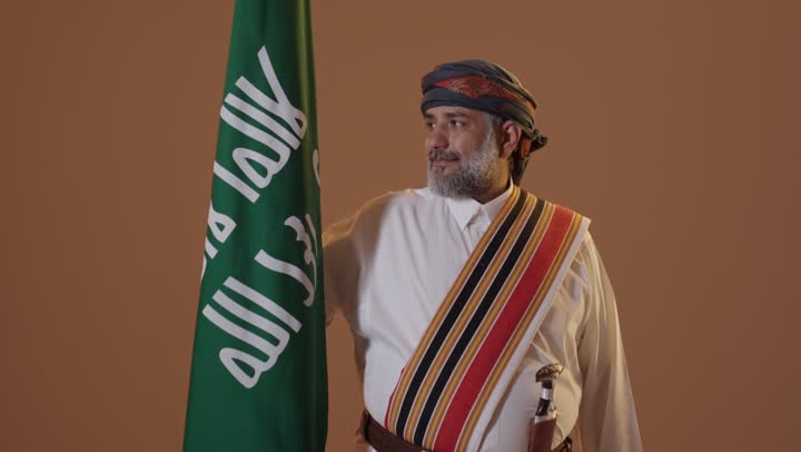 الفخر والاعتزاز بإنجازات الوطن، ذكرى تأسيس الدولة السعودية الأولى 22 فبراير، ارتداء أزياء المنطقة الجنوبية للاحتفال في المناسبات الوطنية،  يوم بدينا 1727م، بورتريه  لرجل كبير بالسن عربي خليجي سعودي  يرتدي الثوب والغترة مع الشال الجنوبي يحمل بيده علم التأسيس السعودي ،  خلفية بيج