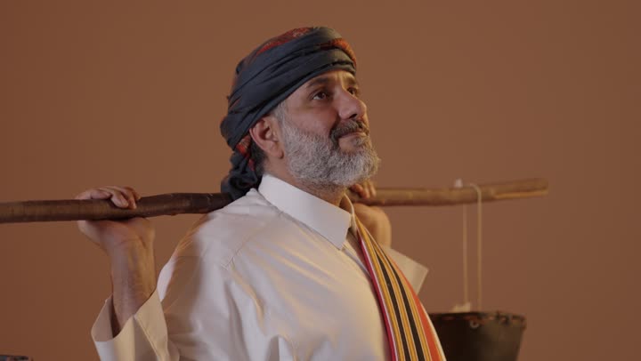 The water bearer is wandering to water people from the well, wearing the attire of the southern region, the establishment of the first Saudi state on February 22, the day we began in 1727 AD, a portrait of an elderly Arab Gulf Saudi man wearing a thobe, ghutrah, and southern shawl carrying a leather bucket, beige background.