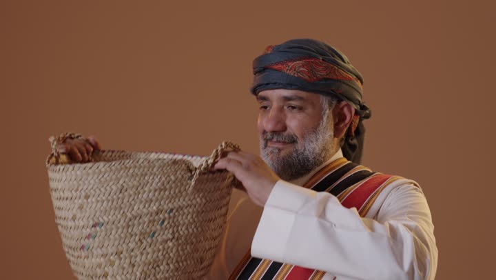 ارتداء أزياء المنطقة الجنوبية،حرف ومهن يدوية قديمة,  ذكرى تأسيس الدولة السعودية 22فبراير، يوم بدينا 1727م،  بورتريه لرجل كبير بالسن عربي خليجي سعودي يرتدي الثوب والغترة مع الشال الجنوبي يضع على كتفه سلة مصنوعة من القش,  خلفية بيج