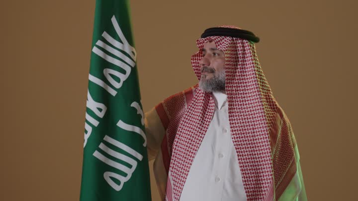 مفهوم حب الوطن والانتماء اليه, يوم بدينا 22 فبراير, ذكرى تأسيس الدولة السعودية الأولى1727م, ارتداء الزي الشعبي في مناسبة وطنية, بورتريه لرجل كبير بالسن عربي خليجي سعودي يرتدي الثوب التقليدي والشماغ يمسك علم يوم التأسيس بتعابير الشموخ والفخر، خلفية بيج