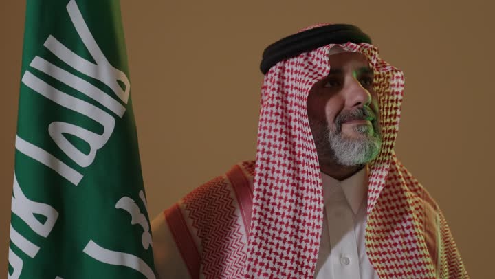 ذكرى تأسيس الدولة السعودية الأولى1727م, يوم بدينا 22 فبراير, ارتداء الزي الشعبي في مناسبة وطنية, مفهوم حب الوطن والانتماء اليه, بورتريه لرجل كبير بالسن عربي خليجي سعودي يرتدي الثوب التقليدي والشماغ يمسك علم يوم التأسيس بتعابير الشموخ والفخر، خلفية بيج
