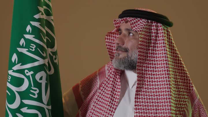 يوم بدينا 22 فبراير, ذكرى تأسيس الدولة السعودية الأولى1727م, ارتداء الزي الشعبي في مناسبة وطنية, مفهوم حب الوطن والانتماء اليه, بورتريه لرجل كبير بالسن عربي خليجي سعودي يرتدي الثوب التقليدي والشماغ يمسك علم يوم التأسيس بتعابير الشموخ والفخر، خلفية بيج