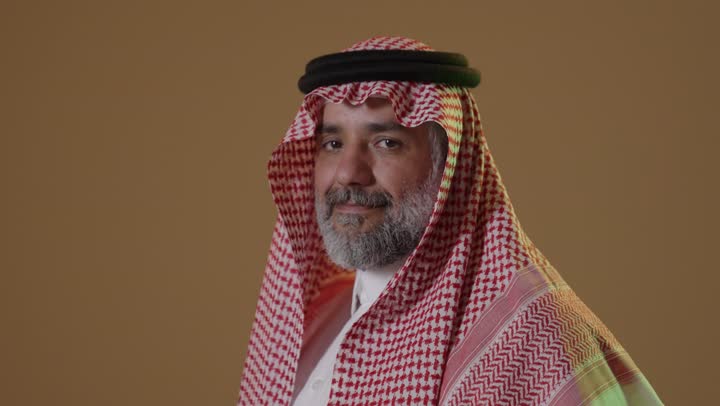 مفهوم الاناقة والرجولة، الإهتمام بالمظهر الخارجي، النظر إلى الكاميرا بايماءات الثقة بالنفس، تعابير الفرح والسعادة، بورتريه لرجل عربي خليجي سعودي يرتدي الثوب التقليدي والشماغ يقف بايدي متقاطعة، خلفية بيج