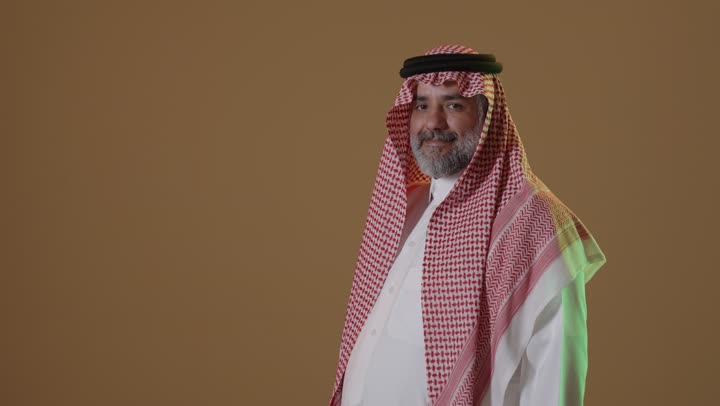 النظر إلى الكاميرا بايماءات الثقة بالنفس، تعابير الفرح والسعادة، مفهوم الاناقة والرجولة، الإهتمام بالمظهر الخارجي، بورتريه لرجل عربي خليجي سعودي يرتدي الثوب التقليدي والشماغ يقف بايدي متقاطعة، خلفية بيج