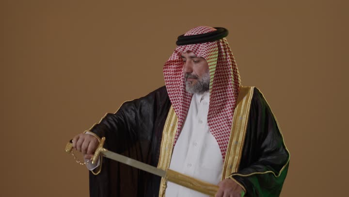 Anniversary of the establishment of the first Saudi state 1727 AD, the concept of love for the homeland and belonging to it, the day we started February 22, wearing traditional attire on a national occasion, a side portrait of an elderly Arab Gulf Saudi man wearing a bisht and agal holding a sword in his hand, beige background.