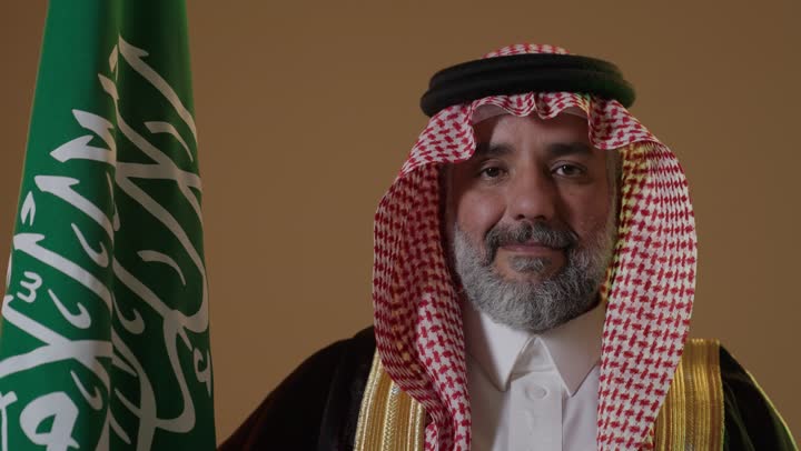 The concept of love for the homeland and belonging to it, the day we started on February 22, the anniversary of the founding of the first Saudi state in 1727, wearing traditional attire on a national occasion, a portrait of an elderly Arab Gulf Saudi man wearing traditional thobe and agal holding the flag of the founding day with expressions of pride and dignity, beige background.