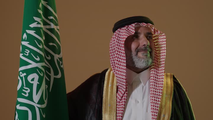 ذكرى تأسيس الدولة السعودية الأولى1727م, مفهوم حب الوطن والانتماء اليه, يوم بدينا 22 فبراير,ارتداء الزي الشعبي في مناسبة وطنية, بورتريه لرجل كبير بالسن عربي خليجي سعودي يرتدي الثوب التقليدي والعقال يمسك علم يوم التأسيس بتعابير الشموخ والفخر، خلفية بيج