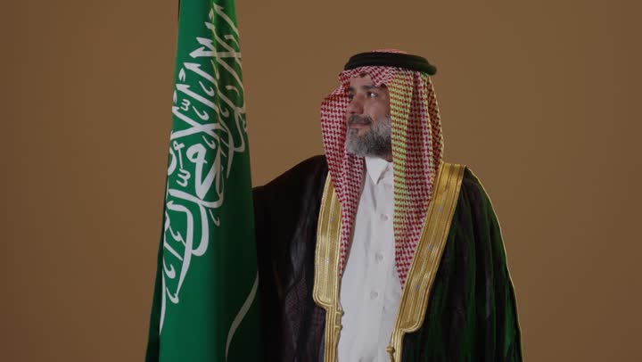مفهوم حب الوطن والانتماء اليه, يوم بدينا 22 فبراير, ذكرى تأسيس الدولة السعودية الأولى1727م, ارتداء الزي الشعبي في مناسبة وطنية, بورتريه لرجل كبير بالسن عربي خليجي سعودي يرتدي الثوب التقليدي والعقال يمسك علم يوم التأسيس بتعابير الشموخ والفخر، خلفية بيج