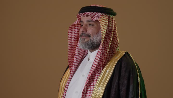 A portrait of an elderly Arab Gulf Saudi man wearing a black bisht and a wide golden agal, holding the flag of Founding Day with expressions of pride and dignity, dressed in traditional attire for a national occasion, embodying the concept of love for the homeland and belonging to it, on the day we began, February 22, the anniversary of the founding of the first Saudi state in 1727, with a beige background.