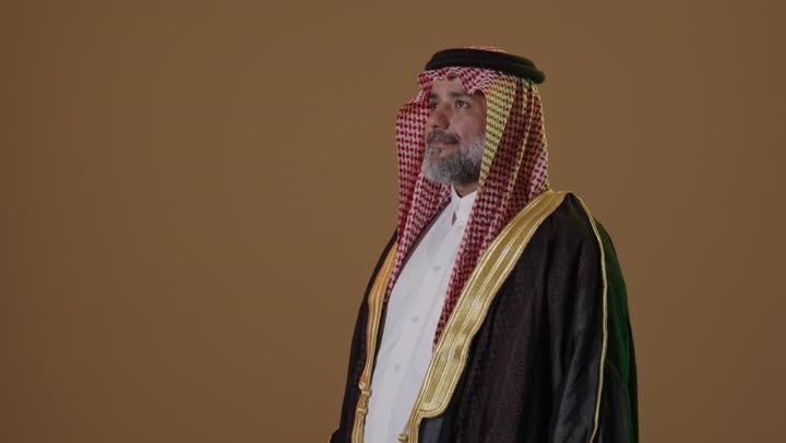 Anniversary of the establishment of the first Saudi state in 1727, wearing traditional attire on a national occasion, the concept of love for the homeland and belonging to it, the day we started, February 22, a portrait of an elderly Arab Gulf Saudi man wearing a black bisht and a wide golden agal holding the flag of the founding day with expressions of pride and dignity, beige background.