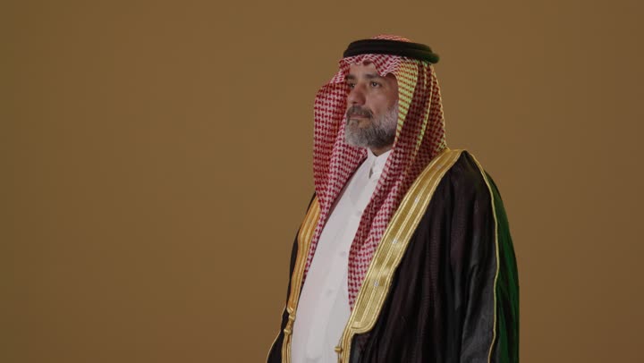 On February 22, we began wearing traditional attire for a national occasion, the concept of love for the homeland and belonging to it, the anniversary of the founding of the first Saudi state in 1727 AD, a portrait of an elderly Arab Gulf Saudi man wearing a black bisht and a wide golden agal holding the flag of the founding day with expressions of pride and dignity, beige background.