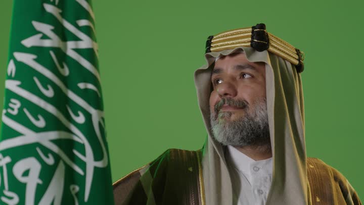 Wearing traditional attire on a national occasion, the concept of love for the homeland and belonging to it, the day we began, February 22, the anniversary of the founding of the first Saudi state in 1727, a portrait of an elderly Arab Gulf Saudi man wearing traditional thobe and agal holding the flag of the founding day with expressions of pride and dignity, green background, chroma.