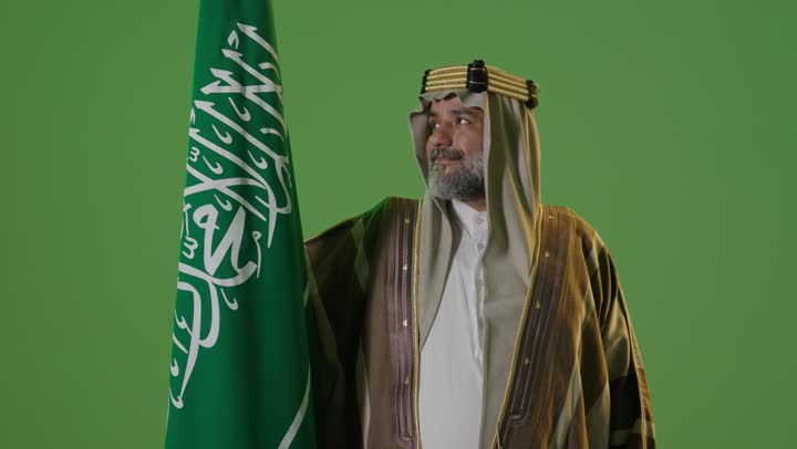 The concept of love for the homeland and belonging to it, the day we started on February 22, the anniversary of the founding of the first Saudi state in 1727, wearing traditional attire on a national occasion, a portrait of an elderly Arab Gulf Saudi man wearing traditional thobe and agal holding the flag of the founding day with expressions of pride and dignity, green background, chroma.