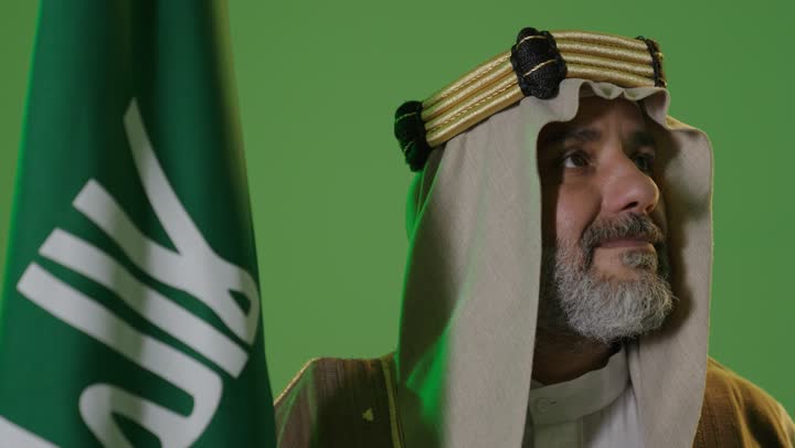 A portrait of an elderly Arab Gulf Saudi man wearing traditional attire and a headband, holding the flag of Founding Day with expressions of pride and dignity, embodying the concept of love for the homeland and belonging to it. The day we began is February 22, the anniversary of the founding of the first Saudi state in 1727. Wearing traditional dress for a national occasion, with a green background, chroma.