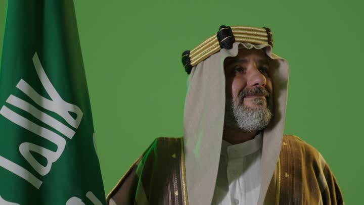 يوم بدينا 22 فبراير, ذكرى تأسيس الدولة السعودية الأولى1727م, مفهوم حب الوطن والانتماء اليه, ارتداء الزي الشعبي في مناسبة وطنية, بورتريه  لرجل كبير بالسن عربي خليجي سعودي يرتدي الثوب التقليدي والعقال يمسك علم يوم التأسيس بتعابير الشموخ والفخر، خلفية خضراء، كروما