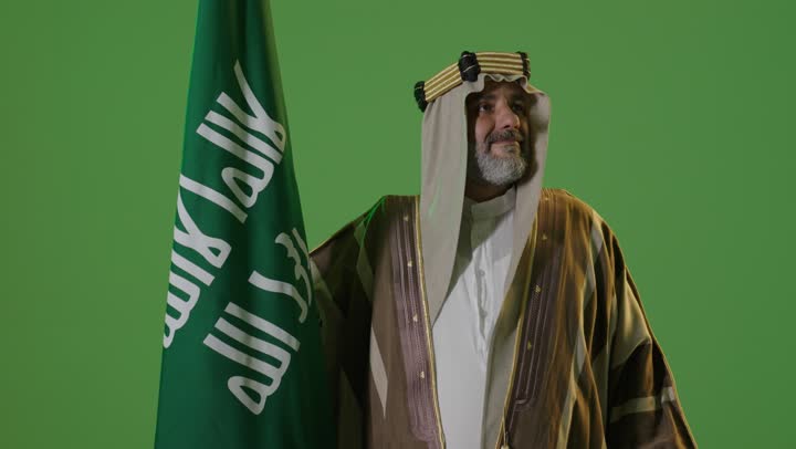 مفهوم حب الوطن والانتماء اليه, يوم بدينا 22 فبراير, ذكرى تأسيس الدولة السعودية الأولى1727م, ارتداء الزي الشعبي في مناسبة وطنية, بورتريه  لرجل كبير بالسن عربي خليجي سعودي يرتدي الثوب التقليدي والعقال يمسك علم يوم التأسيس بتعابير الشموخ والفخر، خلفية خضراء، كروما