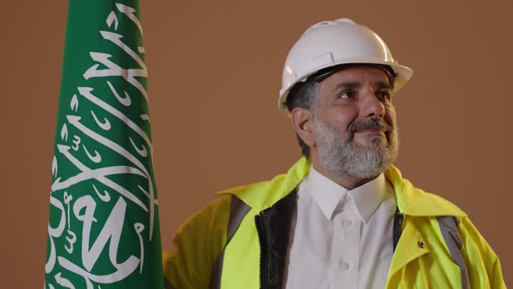 يوم بدينا 22 فبراير, مفهوم حب الوطن والانتماء اليه, ذكرى تأسيس الدولة السعودية الأولى1727م, بورتريه لمهندس كبير بالسن عربي خليجي سعودي يرتديالسترة وخوذة الحماية يمسك علم يوم التأسيس بتعابير الشموخ والفخر، خلفية بيج