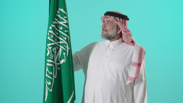 رجل سعودي يرتدي الزي التقليدي مع العلم الوطني السعودي