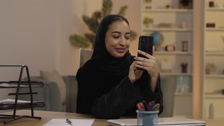 امرأة عربية بالعباءة تستخدم الهاتف الذكي في المكتب المنزلي