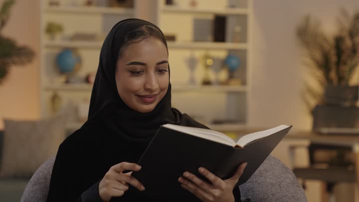 امرأة عربية ترتدي الحجاب تقرأ كتاباً في مكتبة منزلية