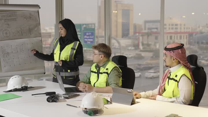 Gender equality in the field of architecture, a Saudi Arabian Gulf Arab woman wearing a black abaya stands next to the architectural plans displayed for explanation and clarification, a Saudi Arabian Gulf Arab man wearing a white thobe and a colored ghutrah listens attentively to the organized explanation, a foreign engineer dressed in casual attire and a safety vest sits with the other team members in the held meeting.