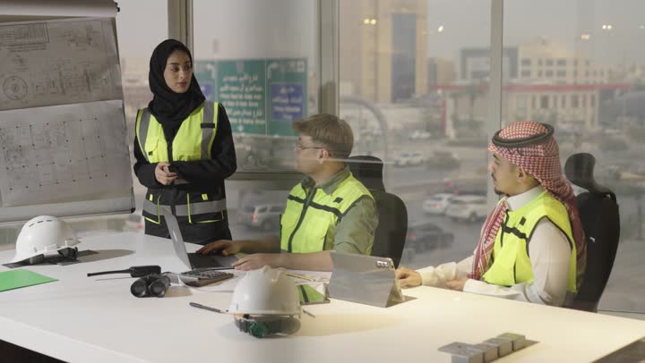 مهندس اجنبي يجلس في الاجتماع مع اعضاء الفريق يشير باصبع السبابة الى المخططات المعلقة , رجل عربي خليجي سعودي يرتدي الثوب الابيض و الشماغ الملون يستمع بانصات الى التوضيح , مهندسة عربية خليجية سعودية ترتدي العباءة السوداء و السترة الواقية تقف بجانب المخططات المعمارية للتوضيح و الشرح , مفهوم العمل الجماعي و المساواة بين الجنسين