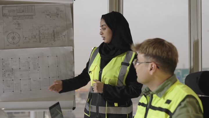 مفهوم الديمقراطية و المساواة داخل المجال الهندسي في المملكة , مهندسة عربية خليجية سعودية محجبة ترتدي العباءة السوداء تقف بجانب المخططات المعلقة , القام بالشرح و التوضيح , مناقشة ودية بين الزملاء للوصول الى القرار الصائب , رجل اجنبي يرتدي نظارة و زي كاجوال يجلس مع باقي الفريق , رجل عربي خليجي سعودي يرتدي الثوب الابيض يشير باصبع السبابة الي الرسومات